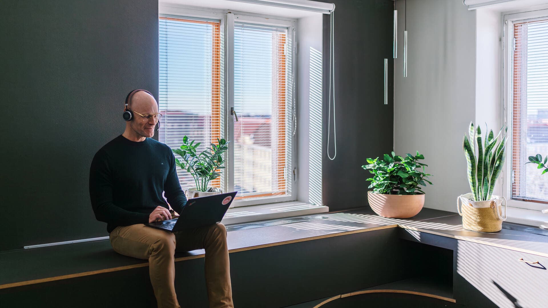Mies istuu neuvotteluhuoneessa kannettava tietokone sylissään, korvakuulokkeet päässään ja katsoo ruudulle hymyillen.