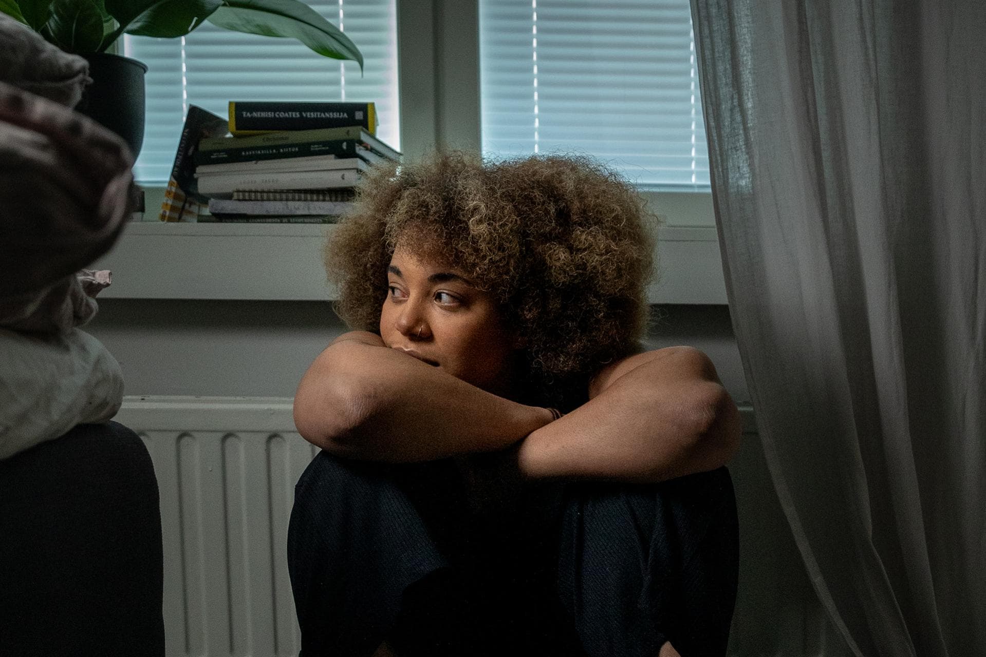 A young woman is sitting in a dark room next to a wall, leaning against her knees.