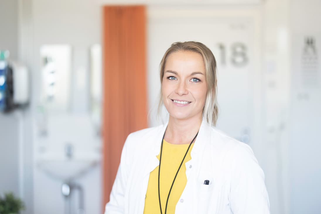 Mehiläisen lääkäri hymyilee iloisesti vastaanottohuoneen oven edessä.