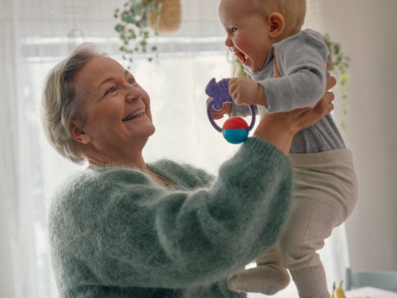 Iäkkäämpi nainen nostaa vauvaa, jolla on leluja käsissään.