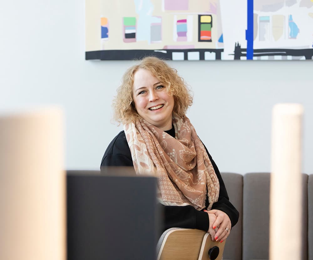 En blonthårig kvinna sitter i soffan och ler. Laura Tero Gofore.