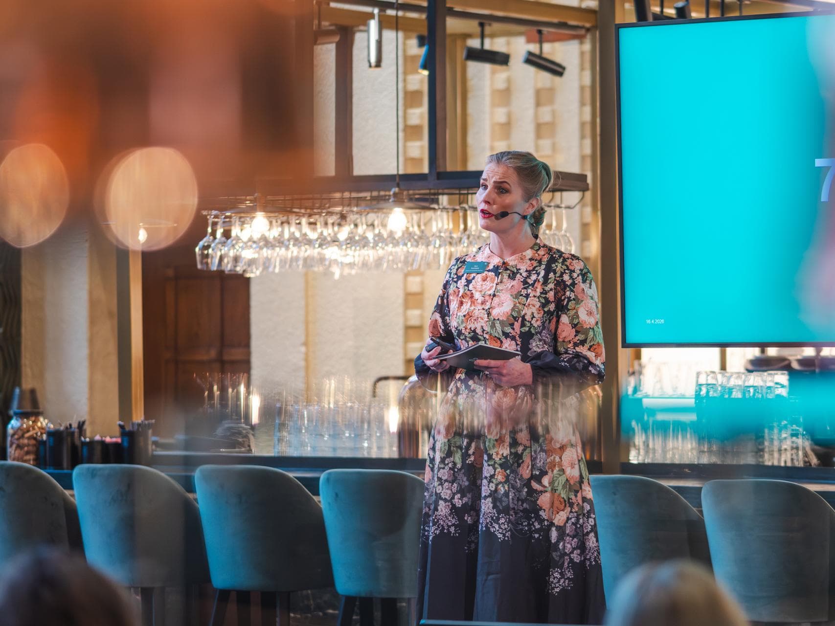 Suvi Suortamo puhuu yleisölle Mehiläisen aamiaistilaisuudessa Ravintola Oliviassa 16.4.2026. Hänellä on yllään kukkakuvioinen mekko ja mikrofoni.
