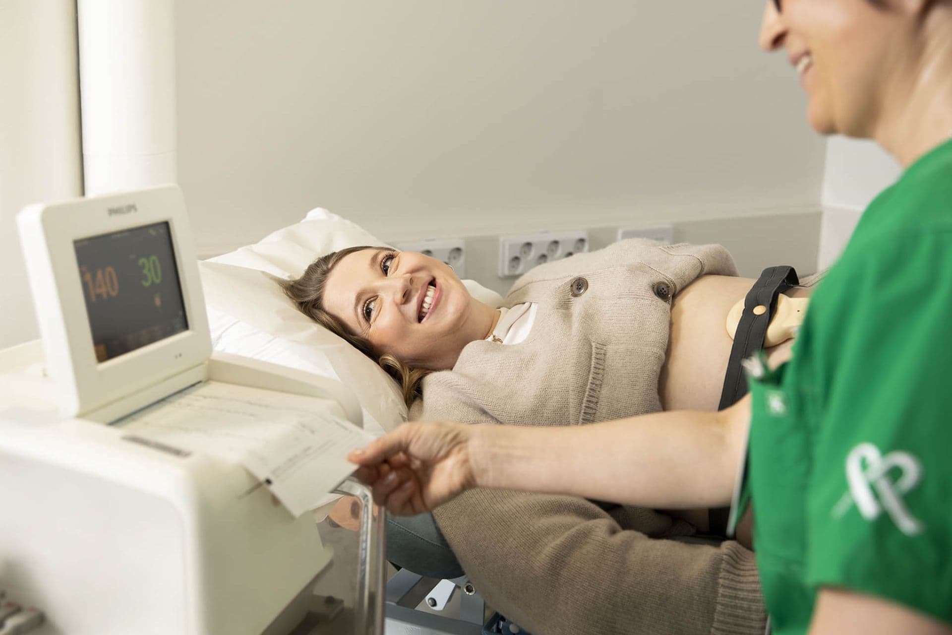 A pregnant woman smiles happily as she lies on the examination table, a fetal monitor attached to her abdomen. A monitor and partially visible healthcare staff are next to her.