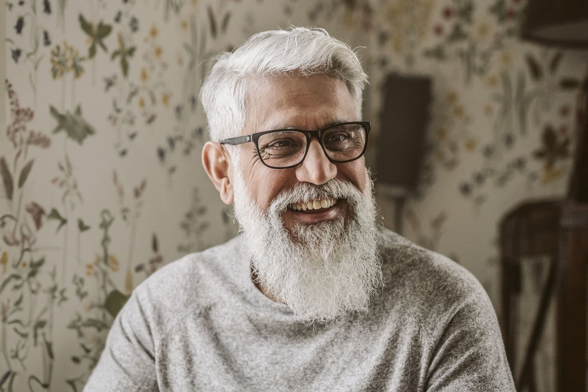 A grey-haired and bearded man smiles happily in his home. His teeth are white and bright.