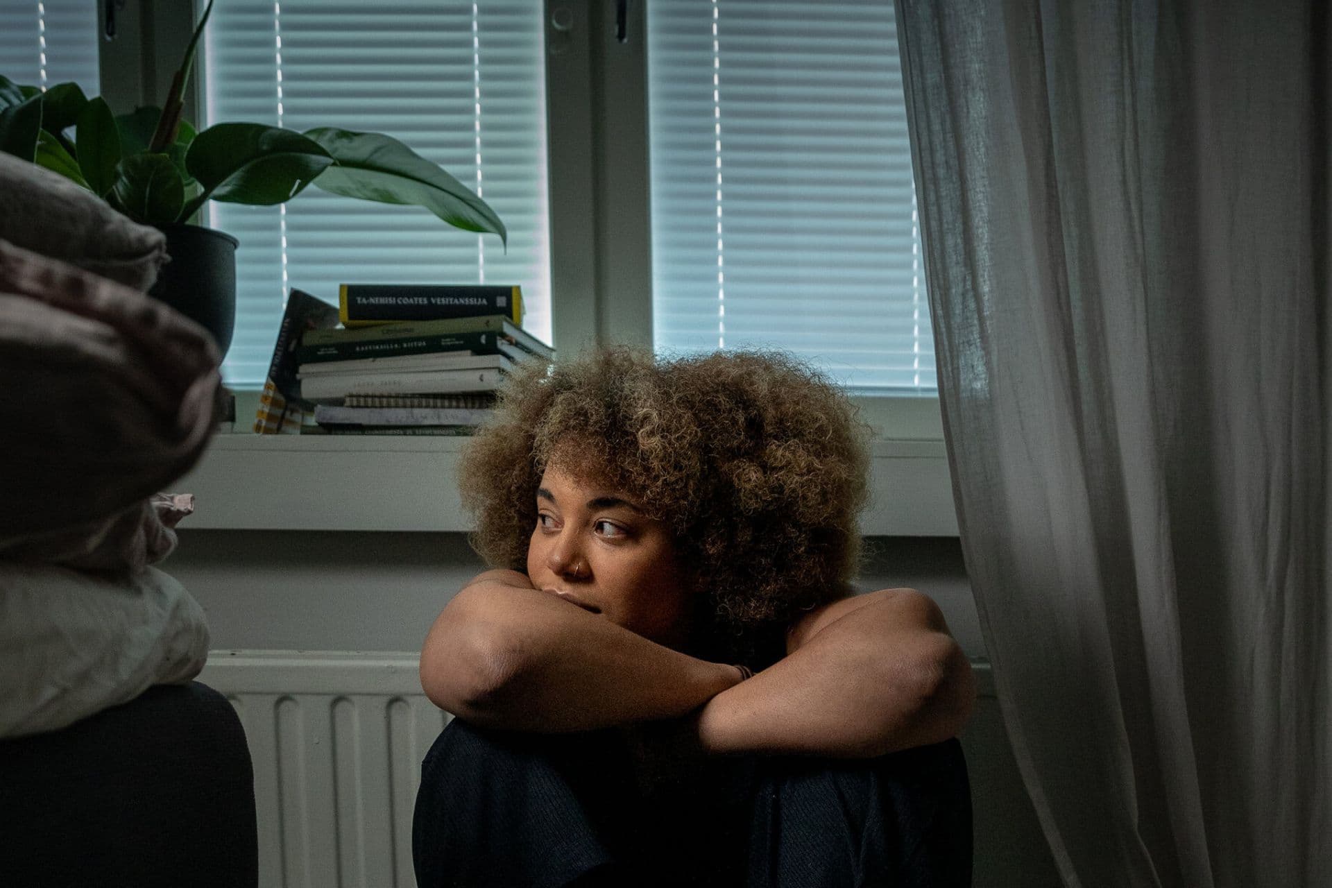 A close-up of a young woman with curly hair and a nose ring. She is sitting indoors and looking away from the camera, with her hands crossed on her knees. In the background, there is a window with blinds and a curtain, and a stack of books on the windowsill.