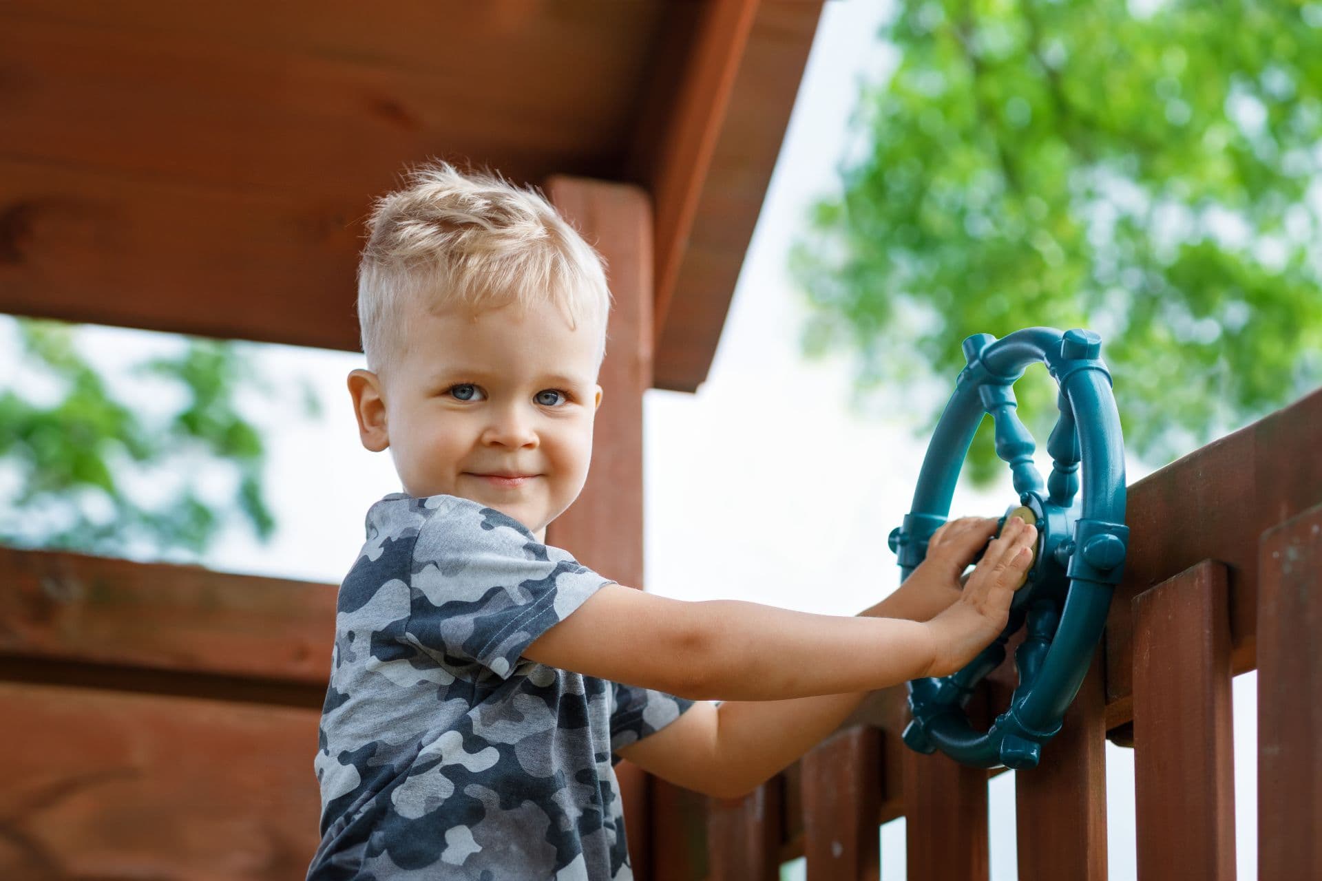 A fair-haired, blue-eyed child smiles in a wooden play structure in a playground, holding onto a turquoise play steering wheel. In the background, green tree leaves and a light sky are visible.
