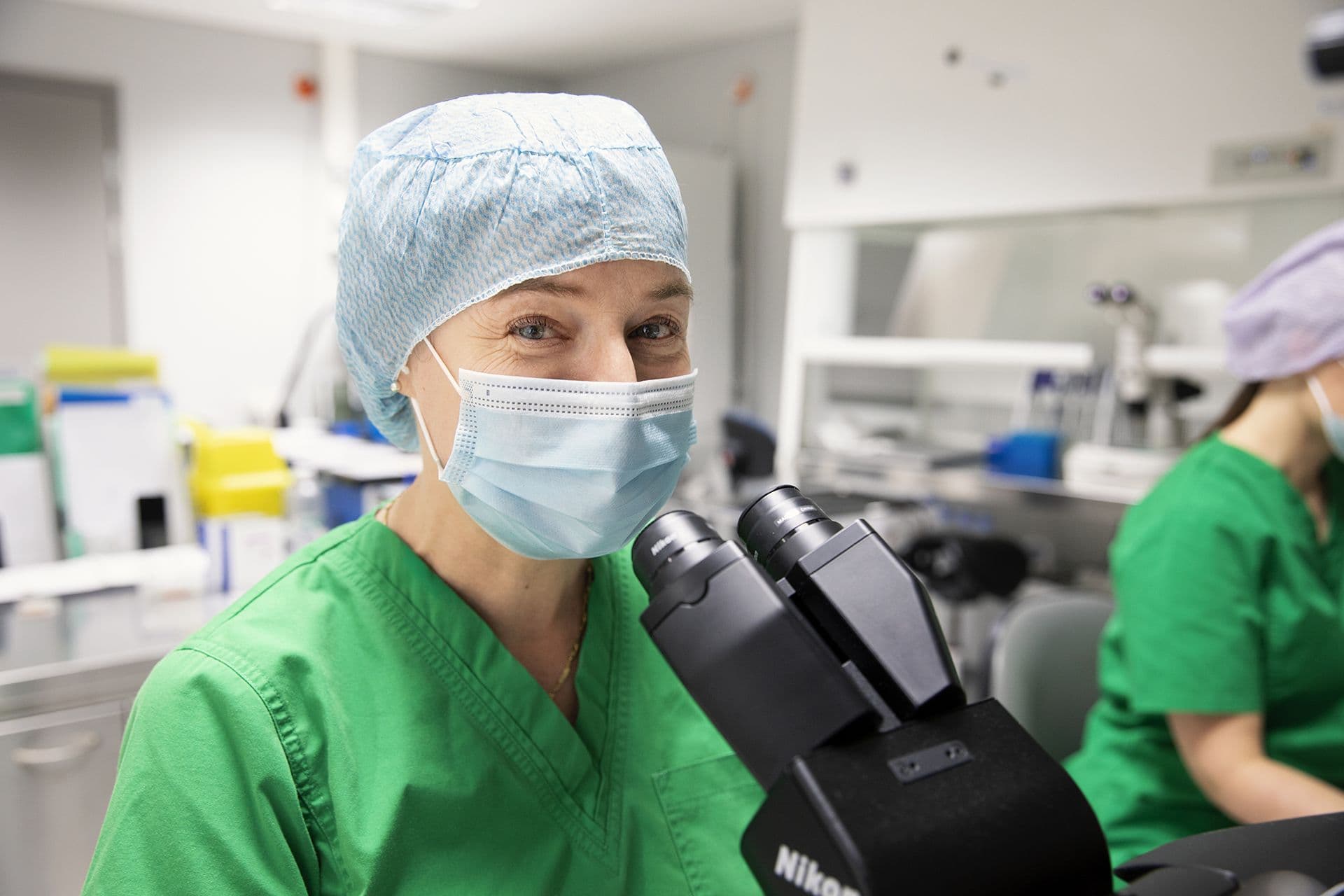 Lääketieteen ammattilainen vihreissä työvaatteissa, suojamyssyssä ja kasvomaskissa katsoo hymyillen kameraan työskennellessään mikroskoopin ääressä laboratoriossa.