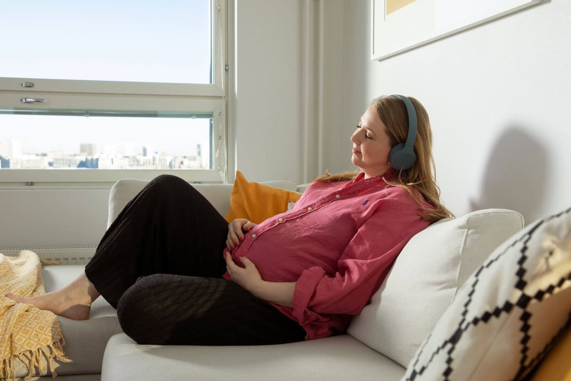 En gravid kvinna ligger på soffan med hörlurar på huvudet och händerna på magen. Hon har på sig en ljusrosa tröja och mörka byxor. I bakgrunden syns ett fönster och ett stadslandskap.