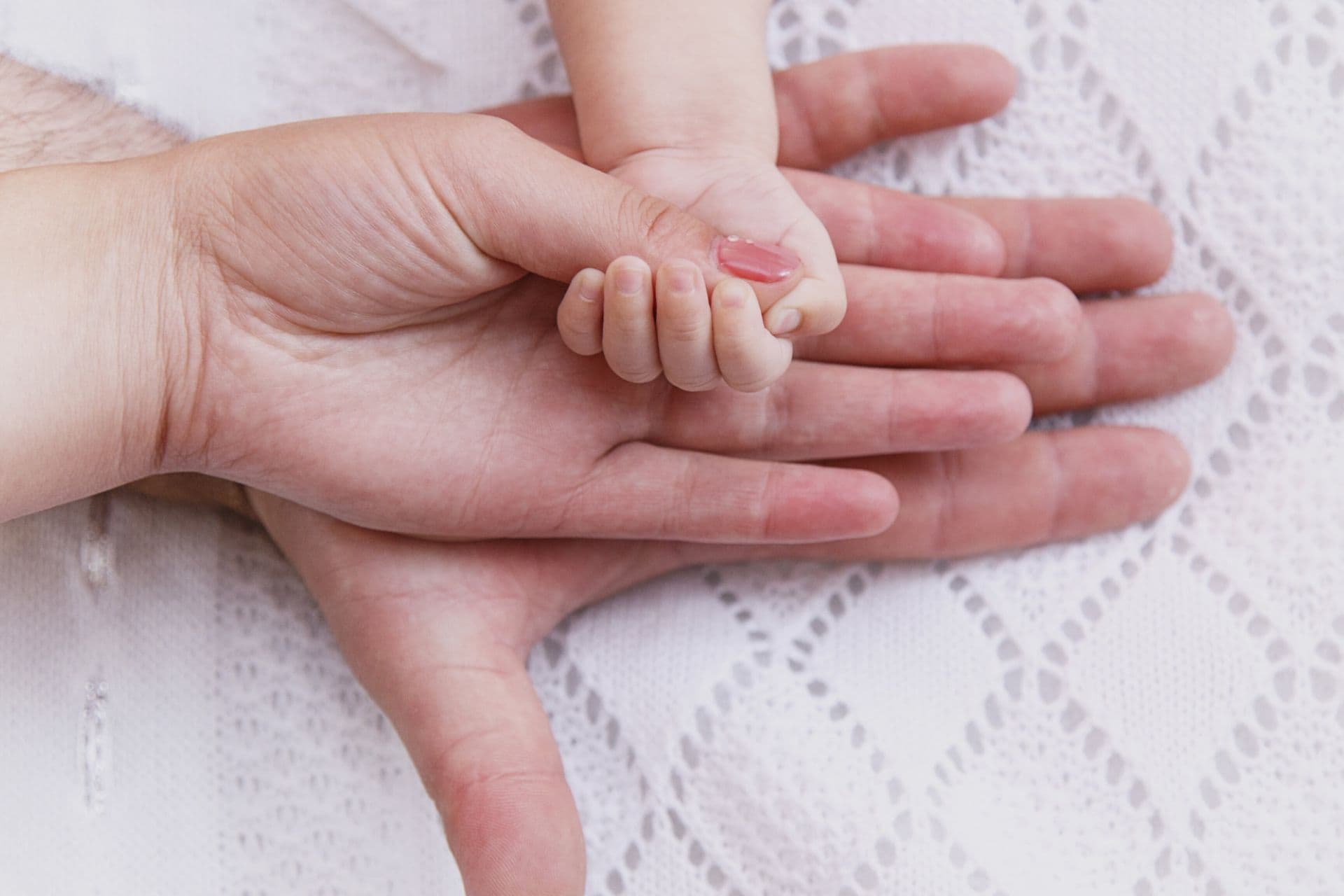 A close-up of three hands stacked on top of each other on a white lace-patterned fabric. At the bottom is an adult man’s hand, on top of it an adult woman’s hand, and on top of all a small baby’s hand grasping the woman’s finger.