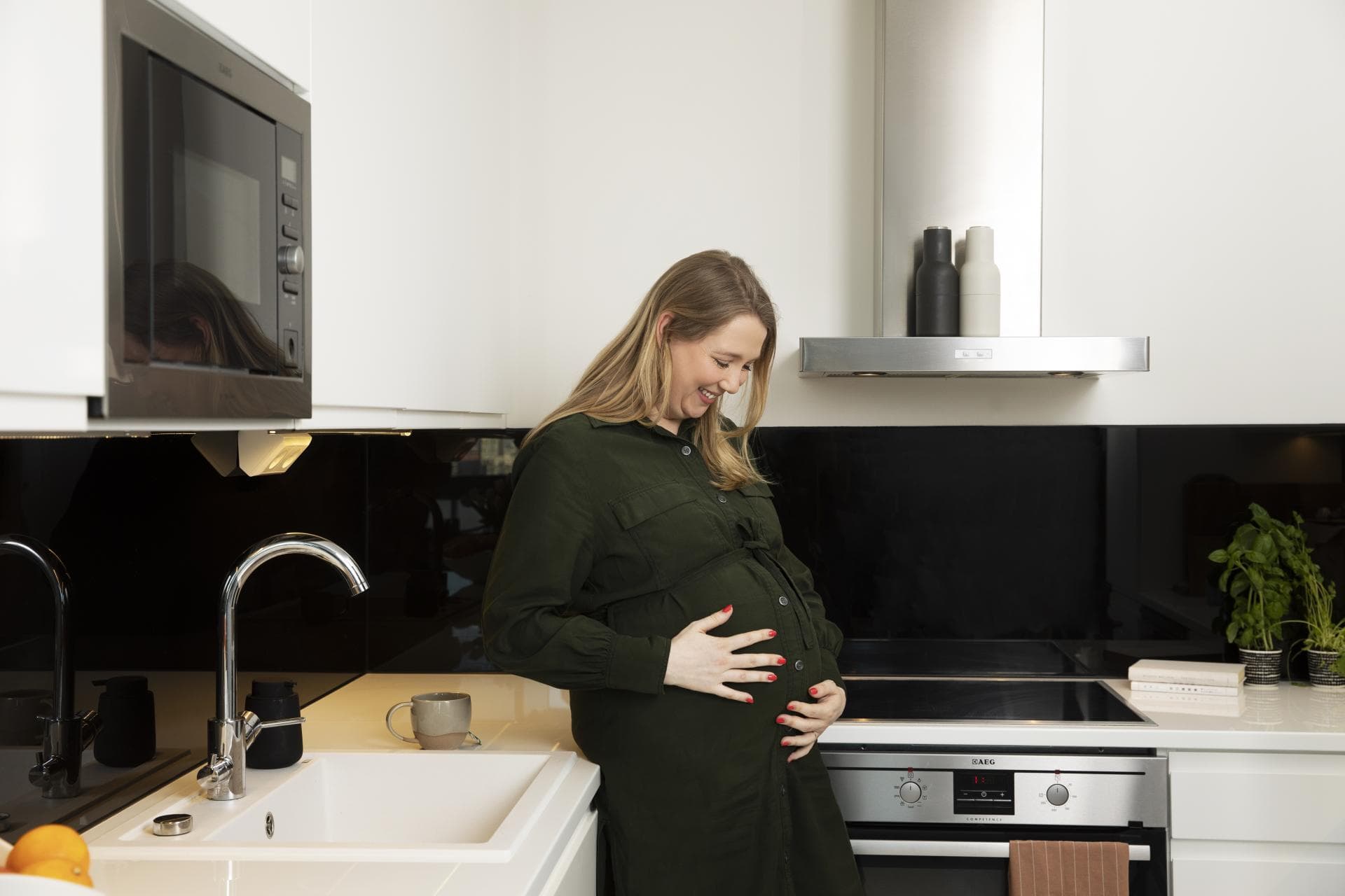 A pregnant woman stands in the kitchen and holds her hands on her belly.
