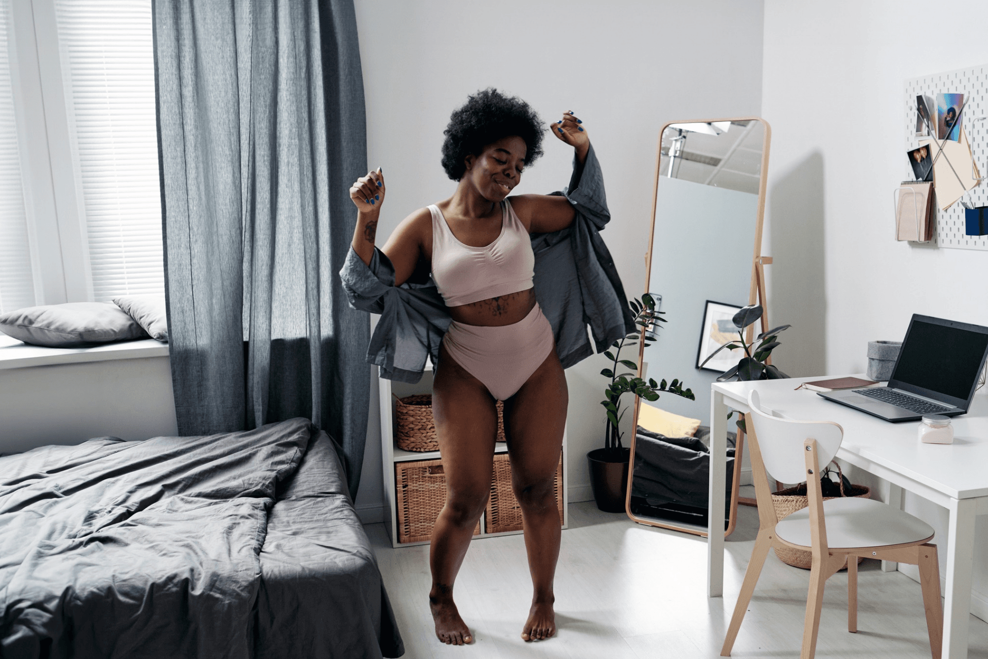 A woman dances casually in her bedroom and smiles contentedly. She is dressed in a sports bra and underpants, and is wearing an open collared shirt.