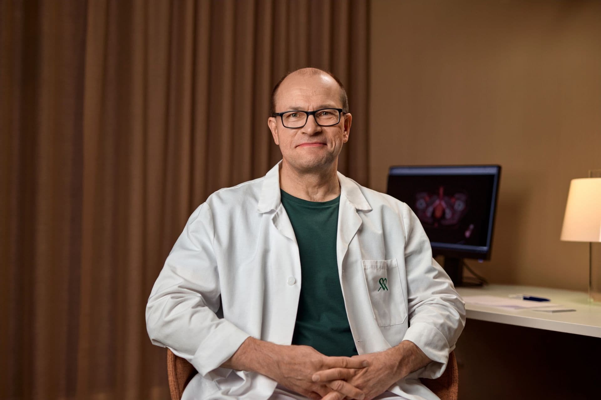 A man in a doctor's coat sits in the consultation room, smiling warmly.