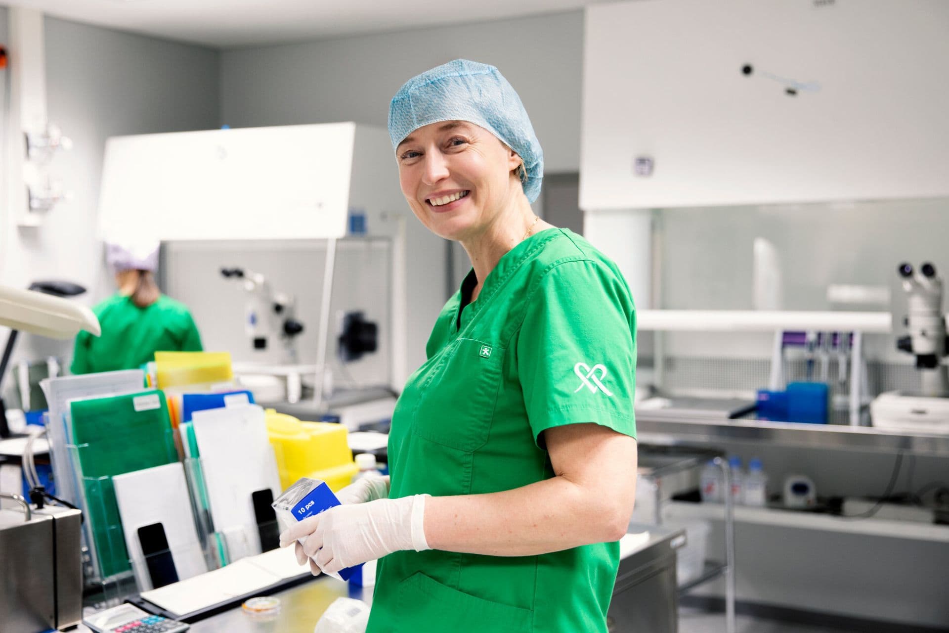 A physician smiles at the camera in a laboratory. She is wearing a green medical coat, white gloves, and a blue hairnet. In the background, laboratory equipment and another person working are visible.