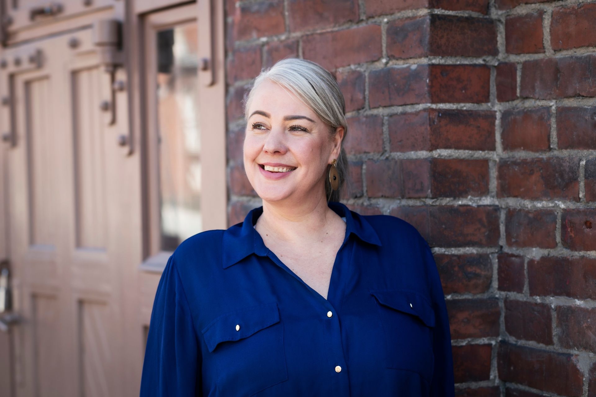 Lähikuva hymyilevästä naisesta, jolla on vaaleat hiukset ja sininen paita. Hän nojaa tiiliseinään ja katsoo vasemmalle.