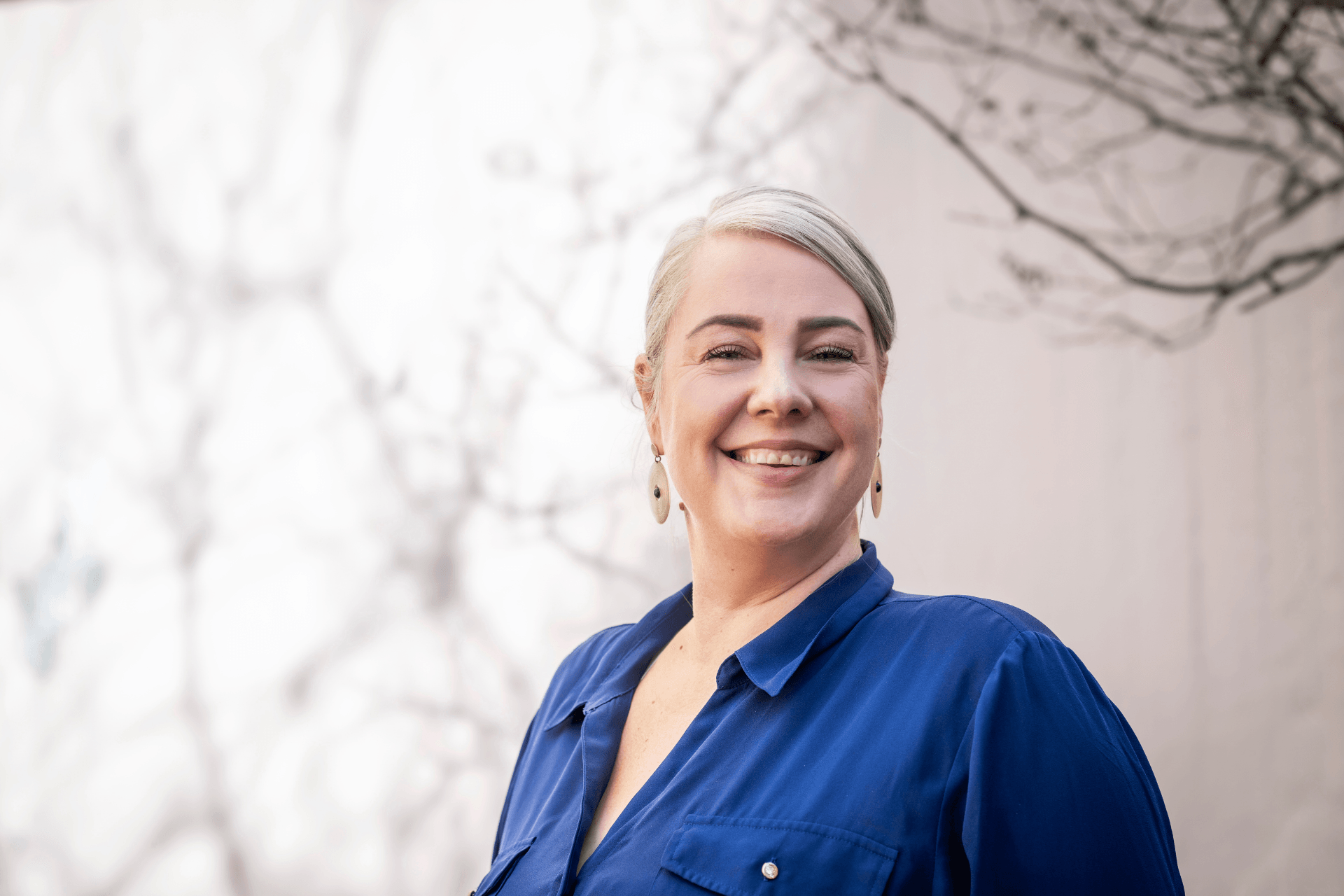 Lähikuva hymyilevästä, keski-ikäisestä naisesta, jolla on vaaleat hiukset ja sininen paita. Hänellä on korvakorut ja taustalla näkyy vaalea tausta ja sumeita oksia.